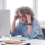 Une femme est assise à un bureau devant un ordinateur, ses mains sont sur les côtés de sa tête et elle a l'air stressée.